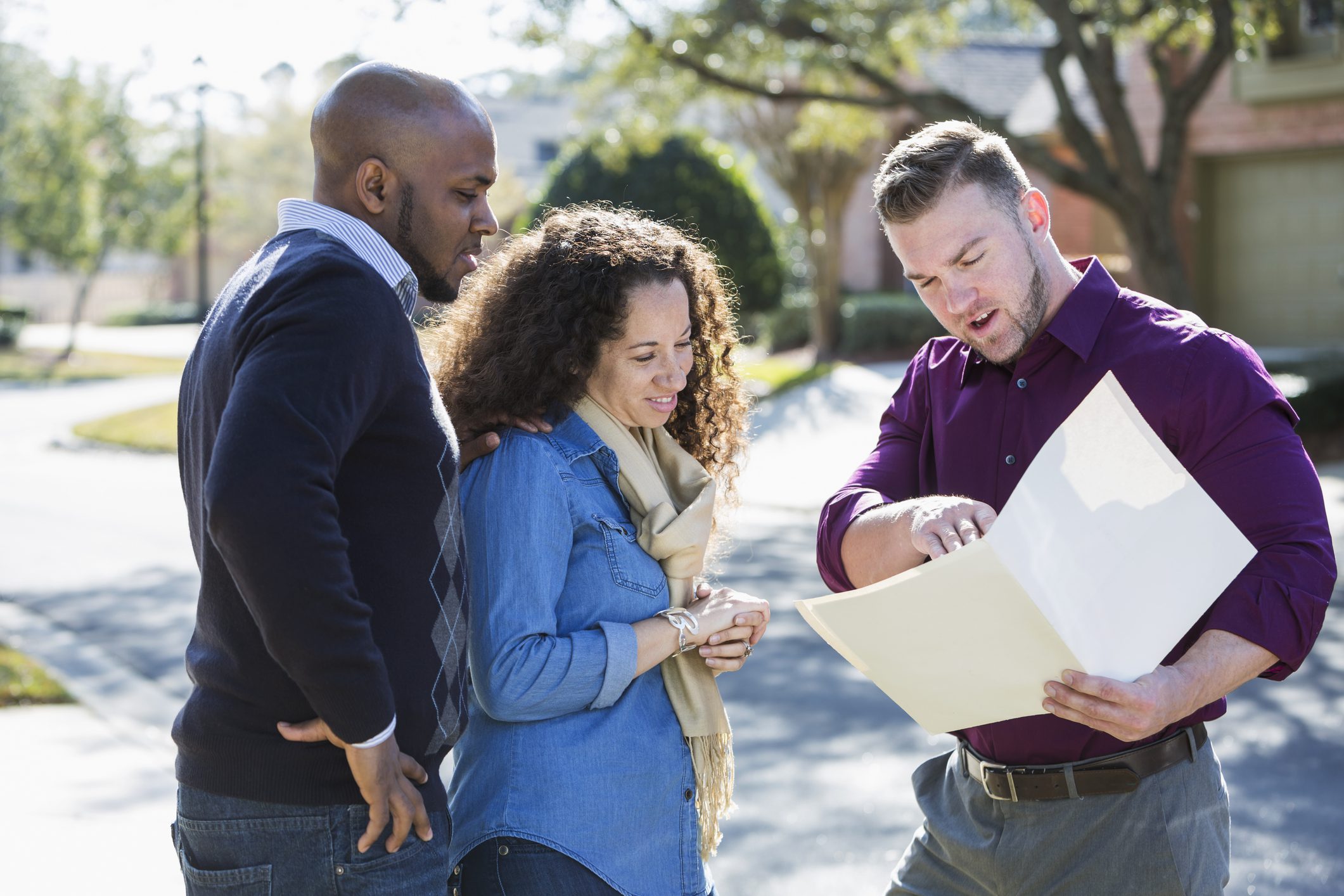 An African American couple standing outdoors on a sunny day on a residential street, with a young man holding a folder.  He is a real estate agent, or maybe a contractor.